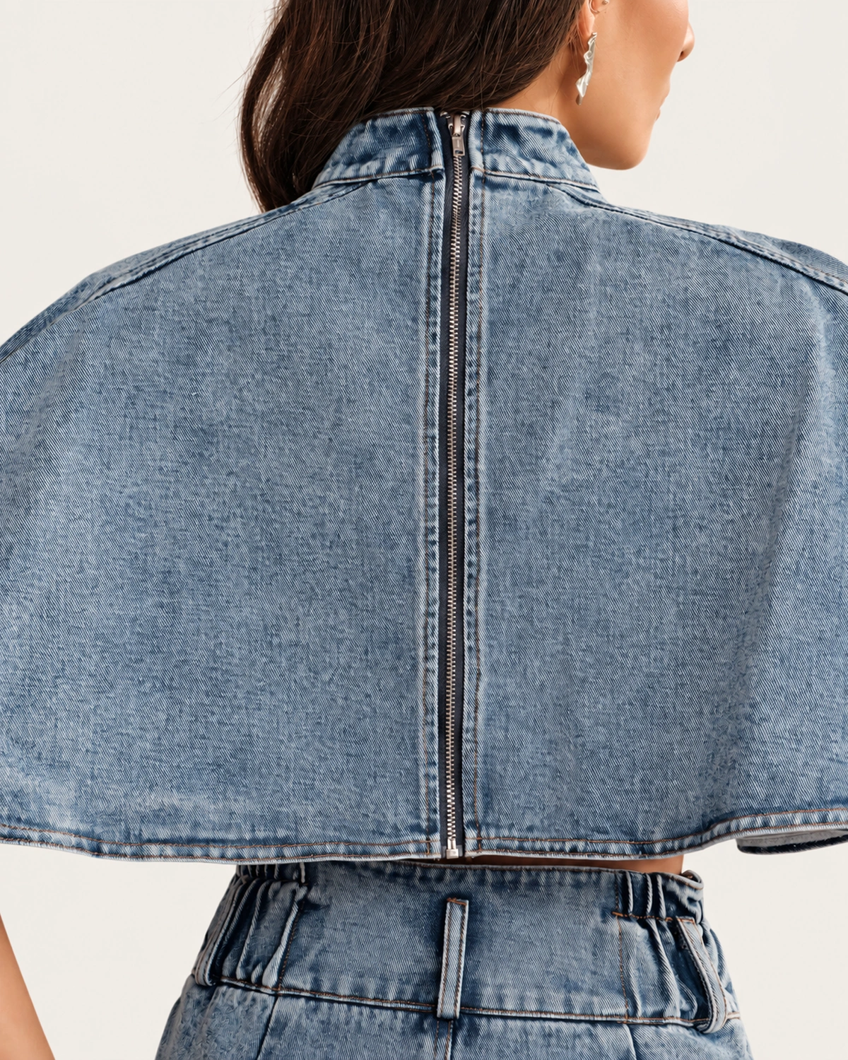 Close posterior da blusa cropped jeans lavagem clara destacando o zíper metálico aparente vestida pela modelo em estúdio mineral