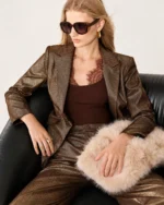 Modelo sentada em poltrona de couro vestindo blazer de alfaiataria bronze metalizado com calça combinando.