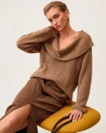 Plano médio da modelo sentada vestindo suéter tricot canelado camel com gola dobrada e saia de camurça.