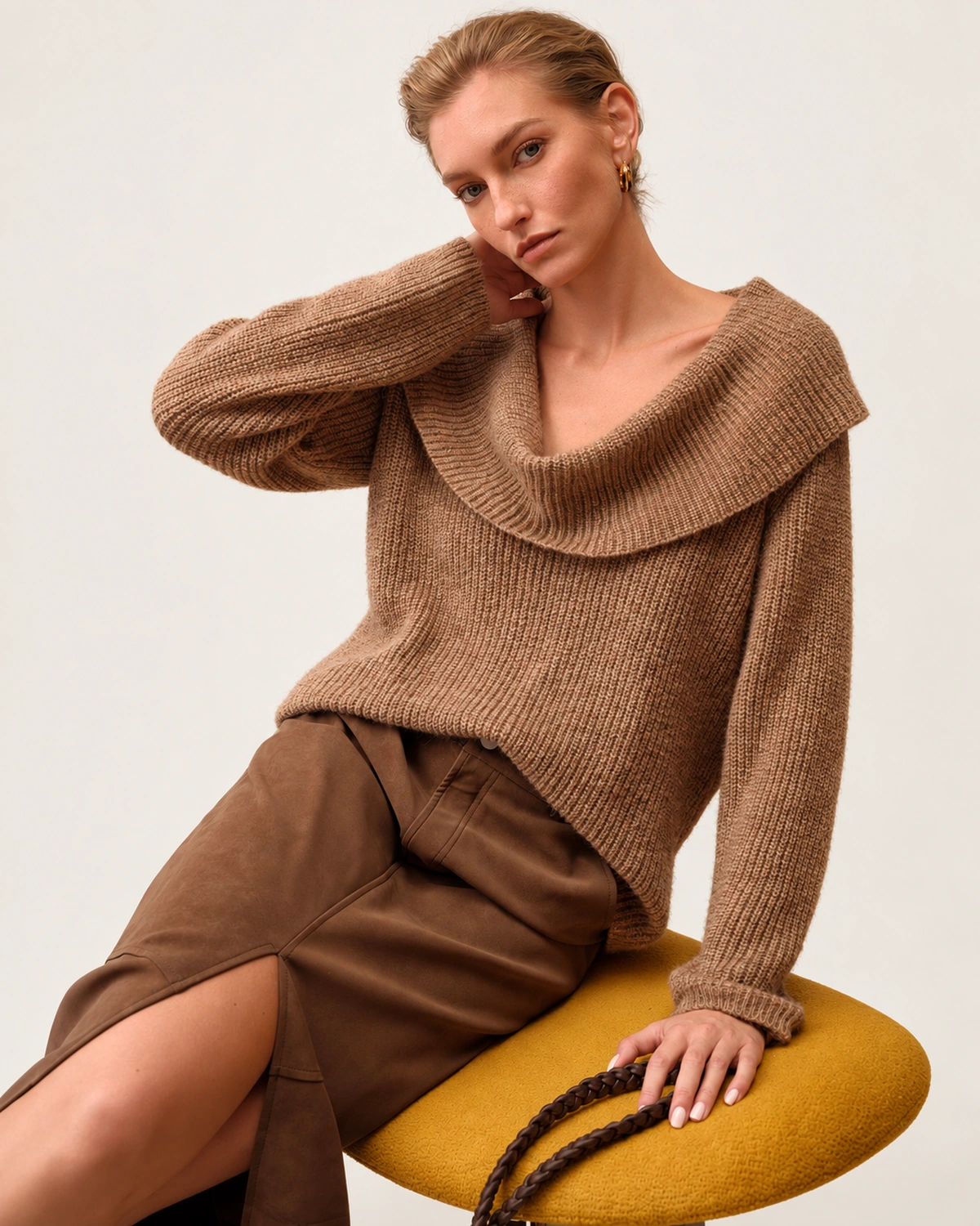 Plano médio da modelo sentada vestindo suéter tricot canelado camel com gola dobrada e saia de camurça.