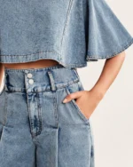 Close de detalhe do fechamento duplo com dois botões metálicos e pregas da calça pantalona jeans clara vestida pela modelo