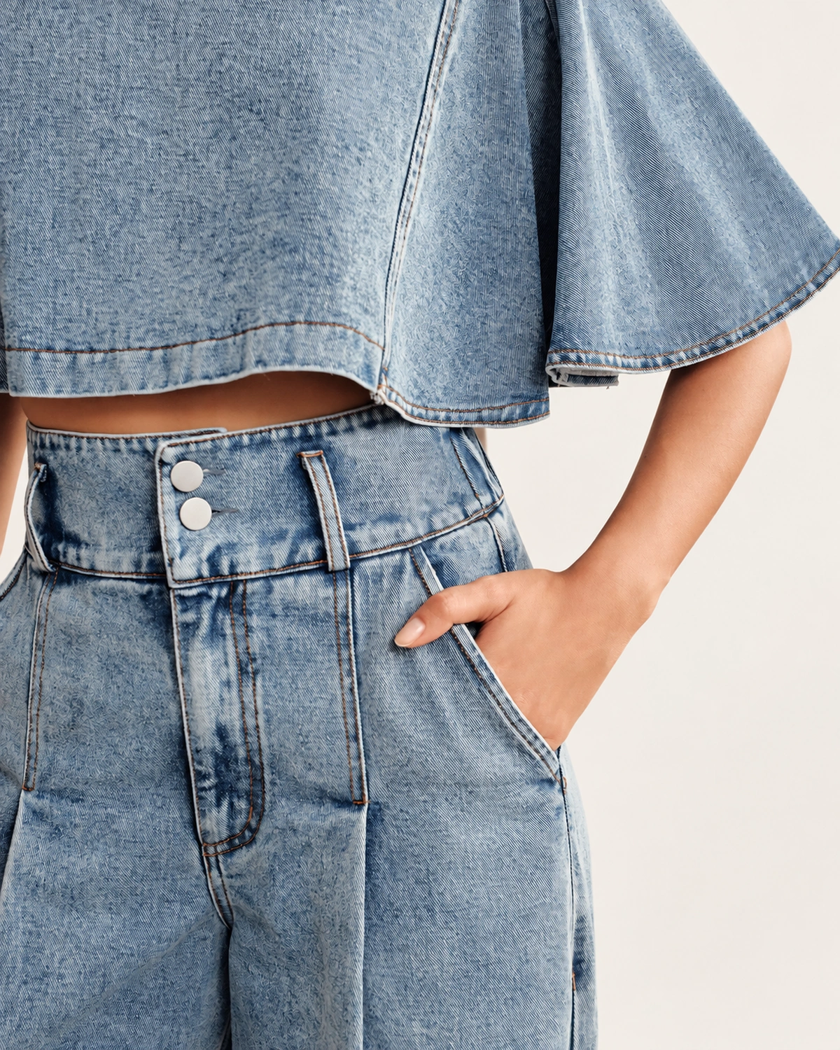 Close de detalhe do fechamento duplo com dois botões metálicos e pregas da calça pantalona jeans clara vestida pela modelo