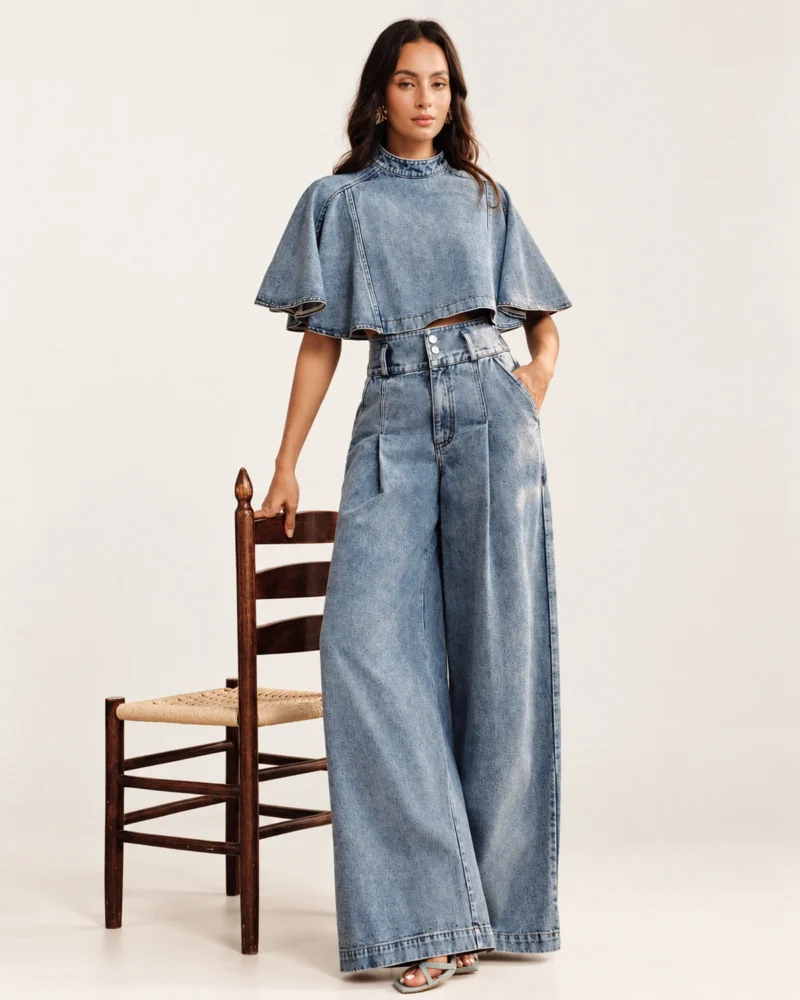 Modelo vestindo conjunto jeans claro blusa evasê e pantalona com mão apoiada em cadeira de estúdio