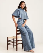 Modelo em ângulo três quartos semi-recostada em cadeira vestindo conjunto jeans claro