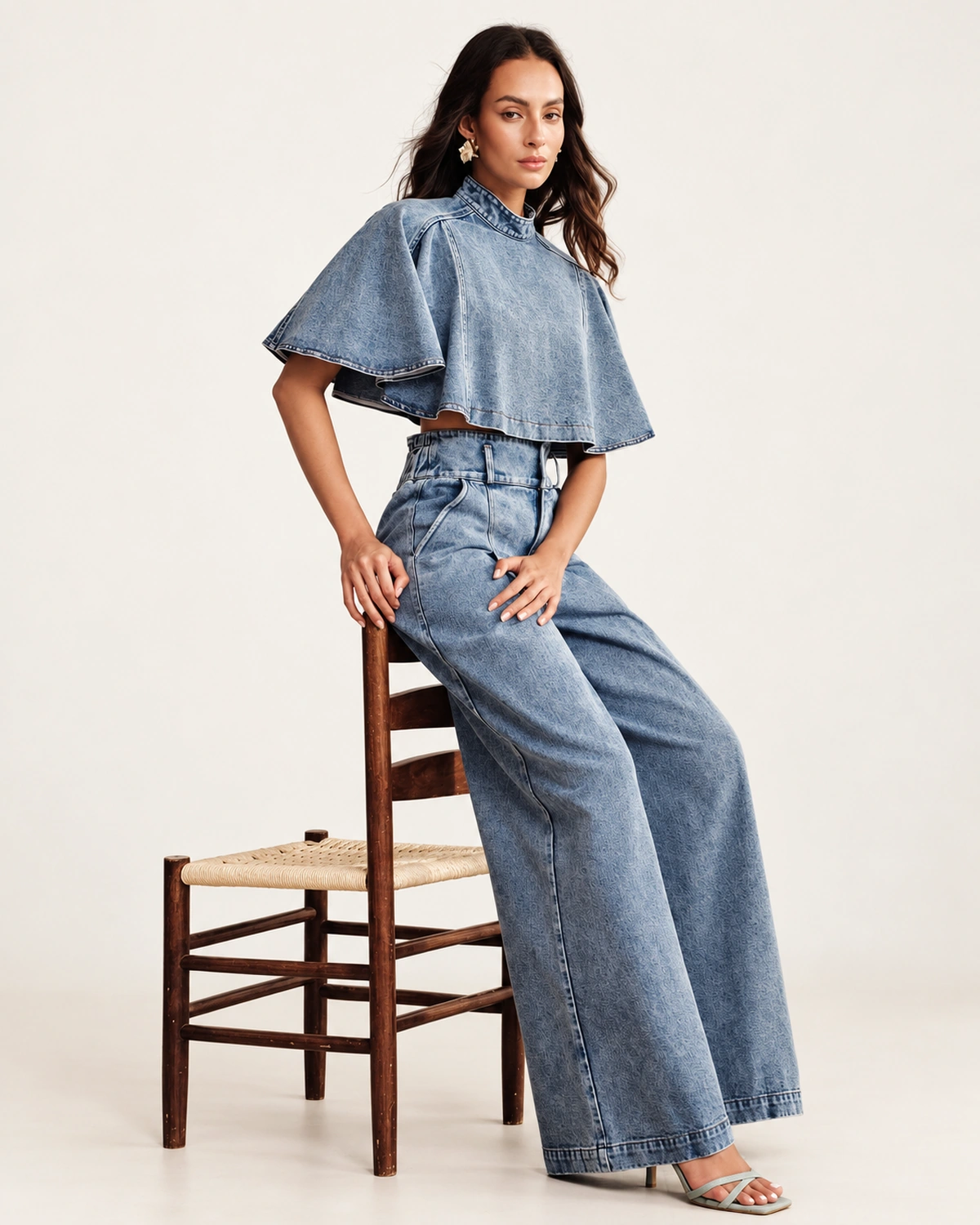 Modelo em ângulo três quartos semi-recostada em cadeira vestindo conjunto jeans claro