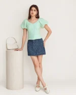 Foto editorial de corpo inteiro da modelo com blusa texturizada Verde-Menta apoiada em coluna de estúdio.