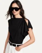 Plano médio da modelo com blusa preta assimétrica, óculos escuros e cinto em estúdio mineral