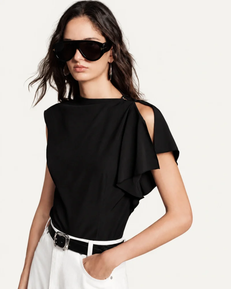 Plano médio da modelo com blusa preta assimétrica, óculos escuros e cinto em estúdio mineral