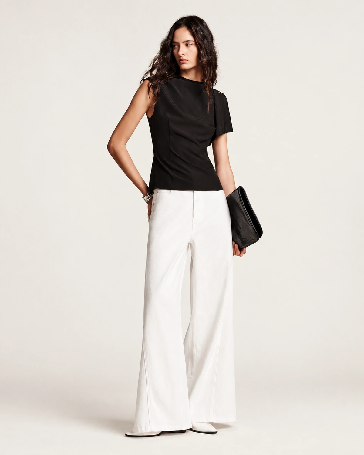 Corpo inteiro da modelo com blusa preta assimétrica, calça pantalona branca e clutch preta