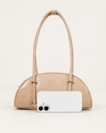 Comparativo de proporção da bolsa meia-lua croco areia ao lado de smartphone no solo mineral.