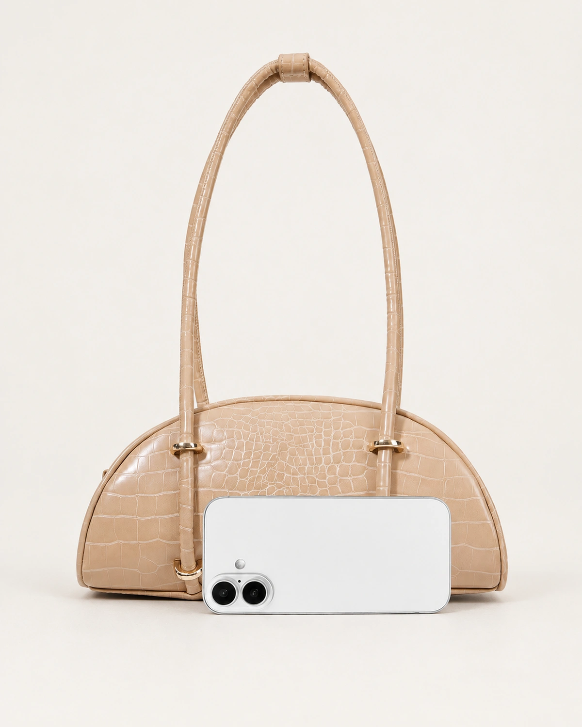Comparativo de proporção da bolsa meia-lua croco areia ao lado de smartphone no solo mineral.