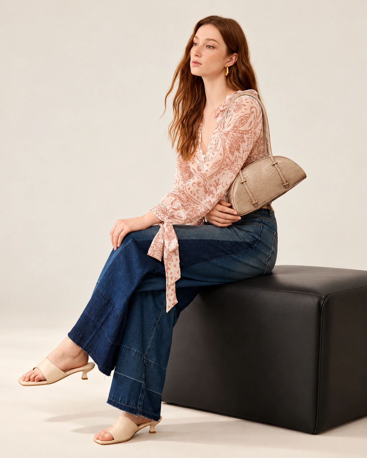 Modelo sentada em banco editorial com a bolsa meia-lua croco areia ao lado.