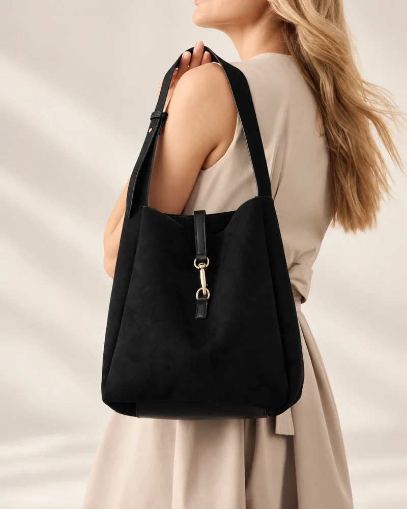 Modelo loira com vestido bege segurando a alça da bolsa shoulder bag preta com detalhe metálico no ombro