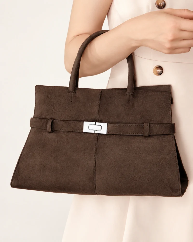 Plano médio da modelo segurando a bolsa tote marrom café no antebraço em look elegante creme de alfaiataria