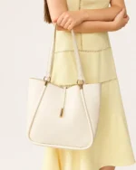 Plano médio da modelo segurando as alças da bolsa tote bege areia à frente do corpo com vestido elegante