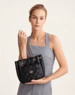 Modelo feminina em conjunto de alfaiataria cinza segurando bolsa tote preta matelassê à frente em estúdio mineral