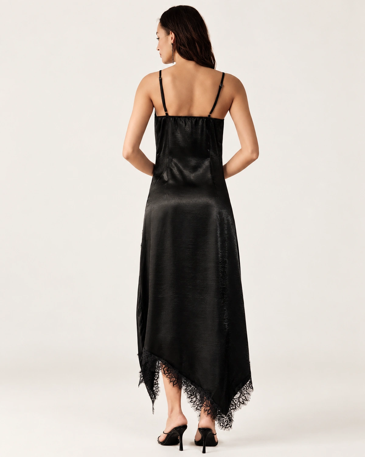 Vista posterior da modelo com vestido slip dress preto acetinado mostrando alças delicadas e decote nas costas.