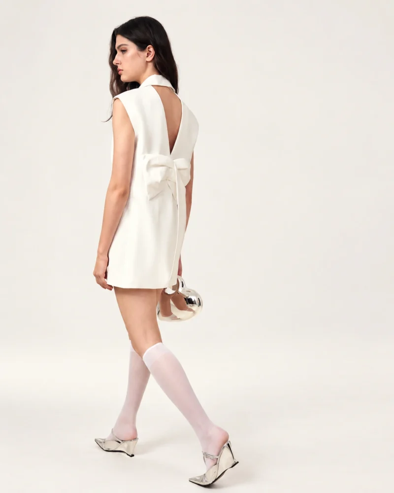 Vista de costas em movimento da modelo exibindo o laço escultural e recorte em V do vestido blazer off-white.