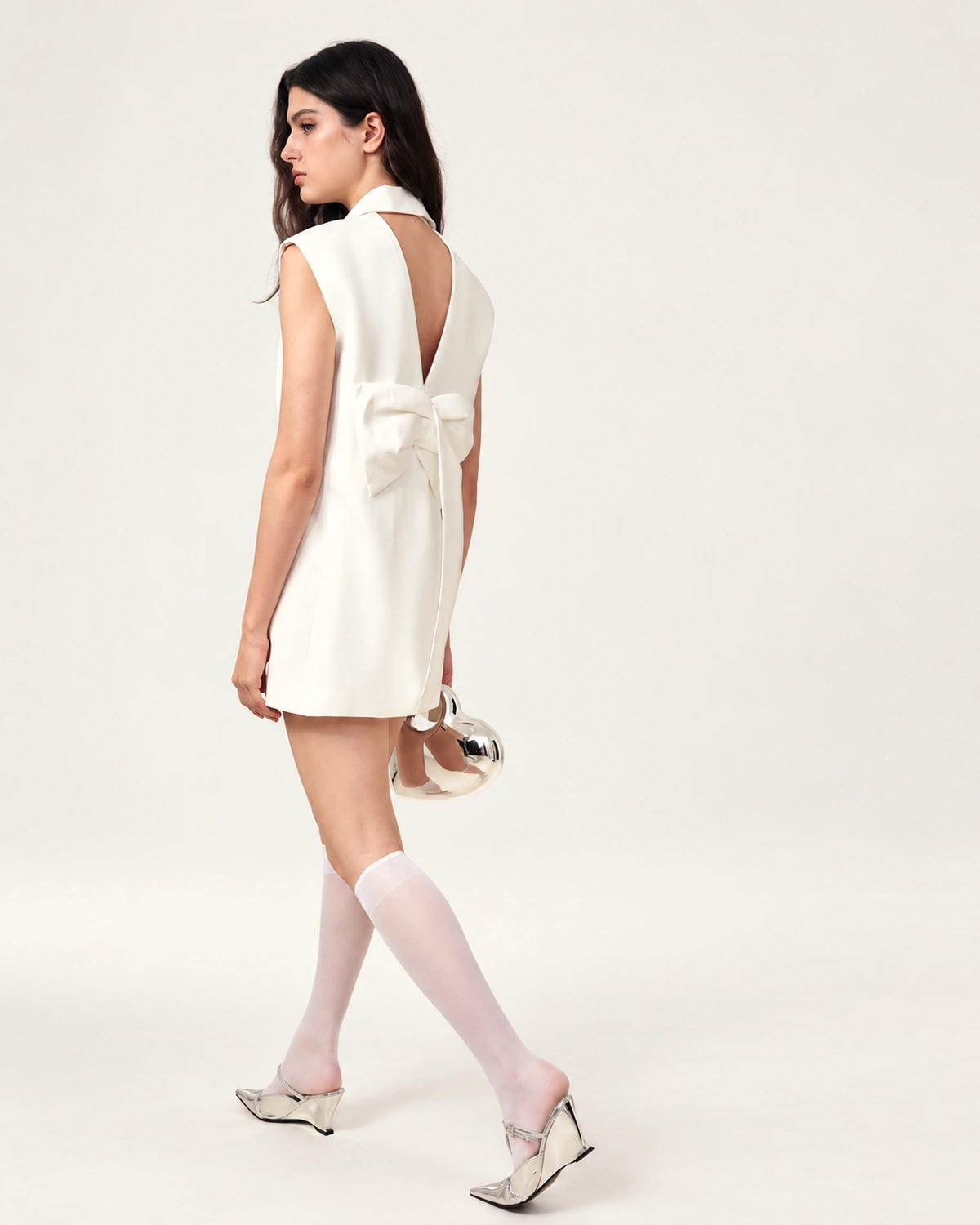 Vista de costas em movimento da modelo exibindo o laço escultural e recorte em V do vestido blazer off-white.