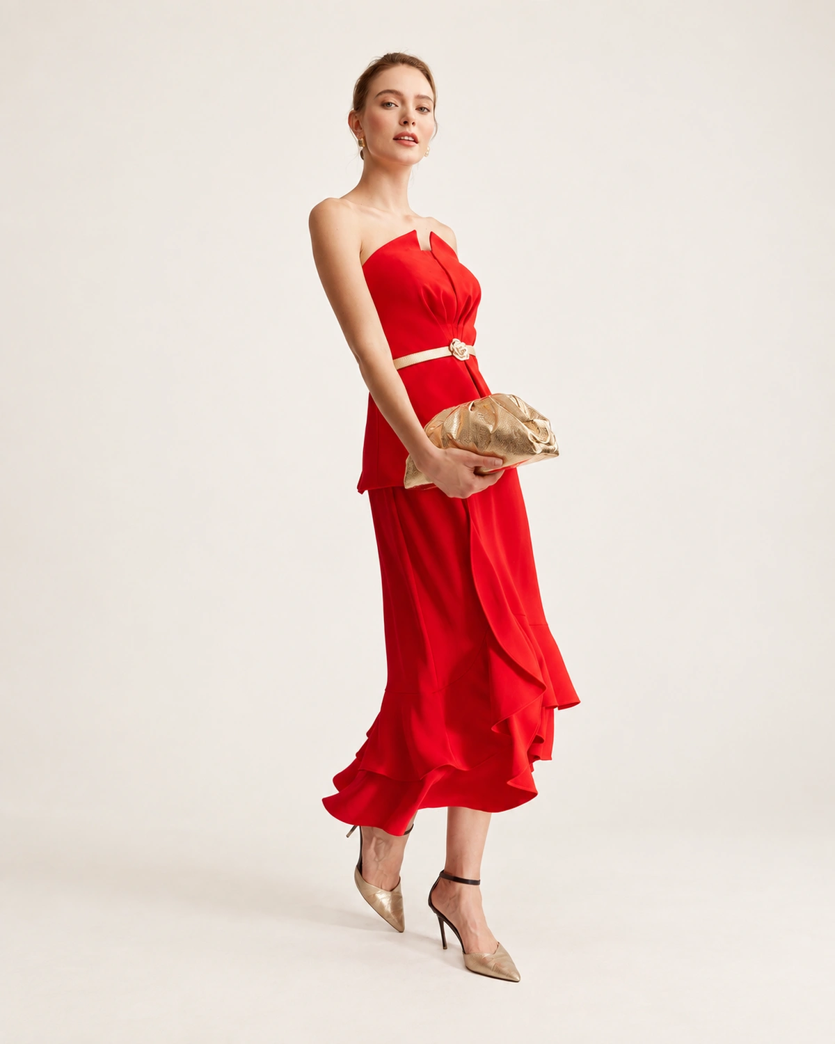 Modelo feminina de corpo inteiro em ângulo três quartos com vestido de festa vermelho e scarpins dourados segurando a clutch na cintura.