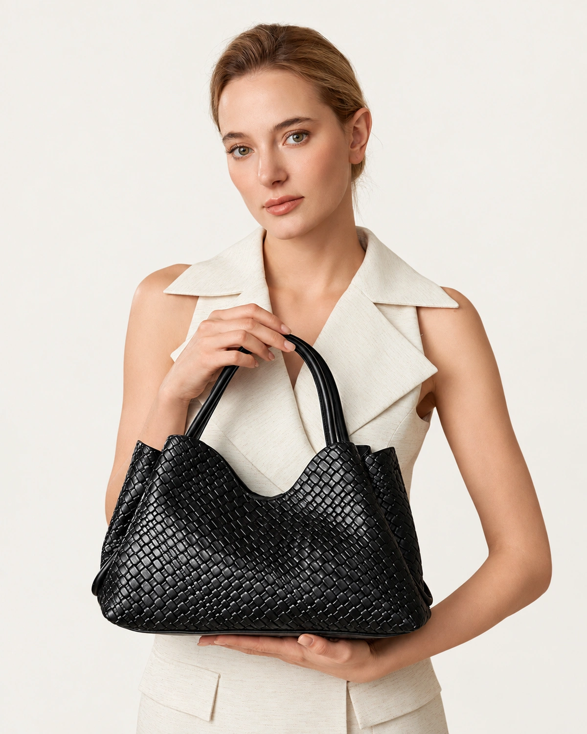 Modelo feminina em plano médio segurando a bolsa tote texturizada preta com ambas as mãos à frente do vestido claro.