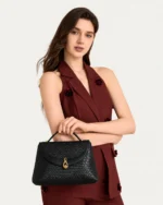 Modelo feminina em pé segurando a Bolsa Satchel Estruturada Tressê preta no estúdio Peaker.