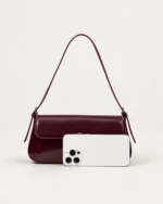 Comparativo de proporção e tamanho real da bolsa tiracolo flap bordeaux ao lado de smartphone no estúdio mineral.
