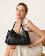 Modelo feminina sentada com vestido claro apoiando a bolsa tote texturizada preta no colo no estúdio mineral.