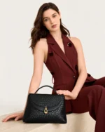 Modelo de alfaiataria bordô sentada ao lado da Bolsa Satchel Estruturada preta com fecho dourado.