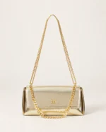 Vista frontal isolada da bolsa tiracolo estruturada com aba flap em couro metalizado dourado acetinado no piso mineral.