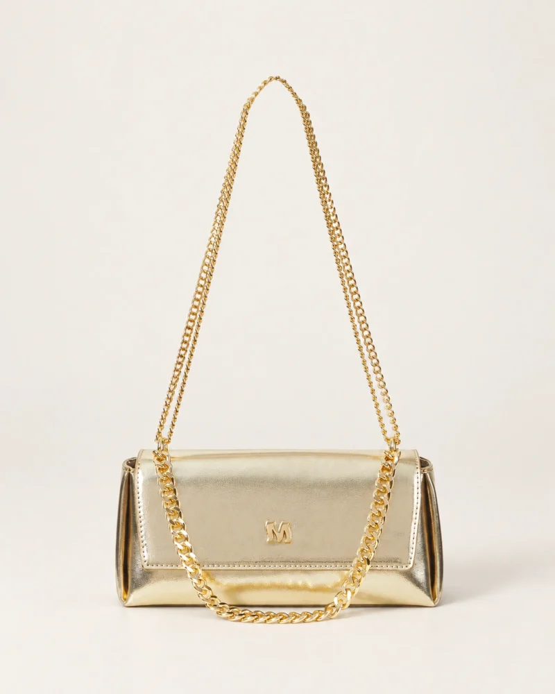 Vista frontal isolada da bolsa tiracolo estruturada com aba flap em couro metalizado dourado acetinado no piso mineral.
