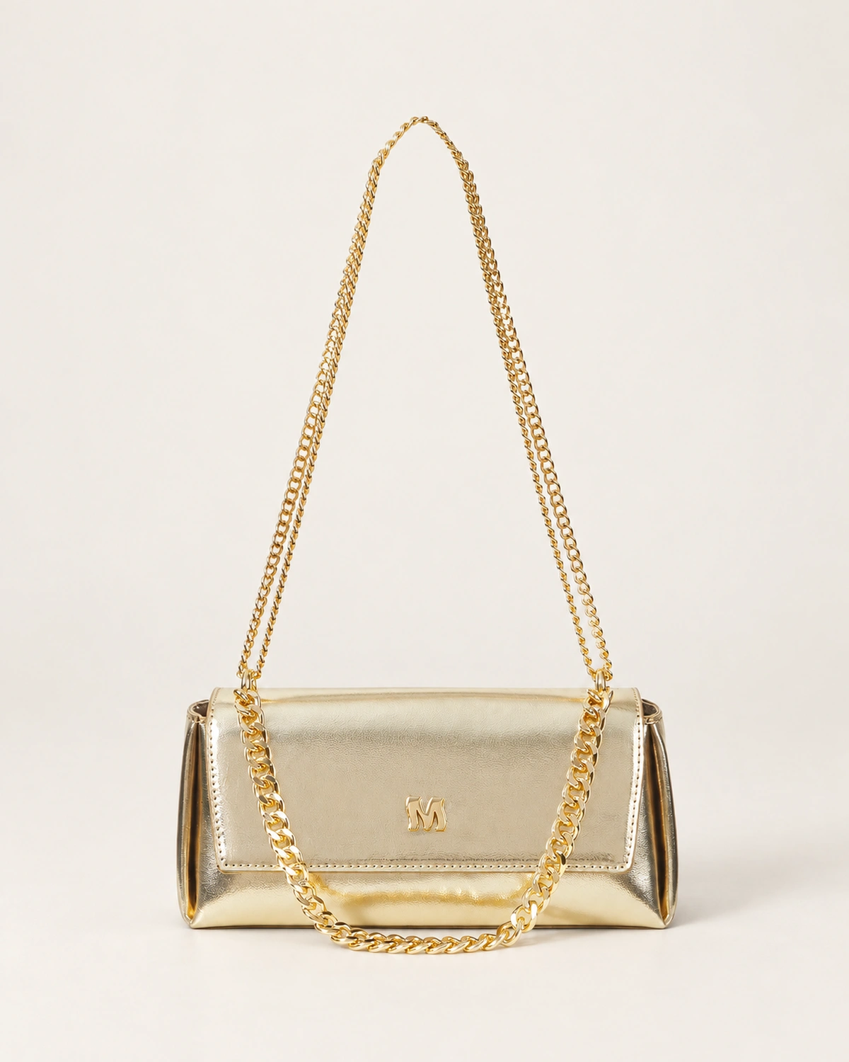 Vista frontal isolada da bolsa tiracolo estruturada com aba flap em couro metalizado dourado acetinado no piso mineral.