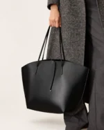Plano detalhe da mão da modelo segurando a alça da bolsa tote preta ao lado da calça de alfaiataria cinza.