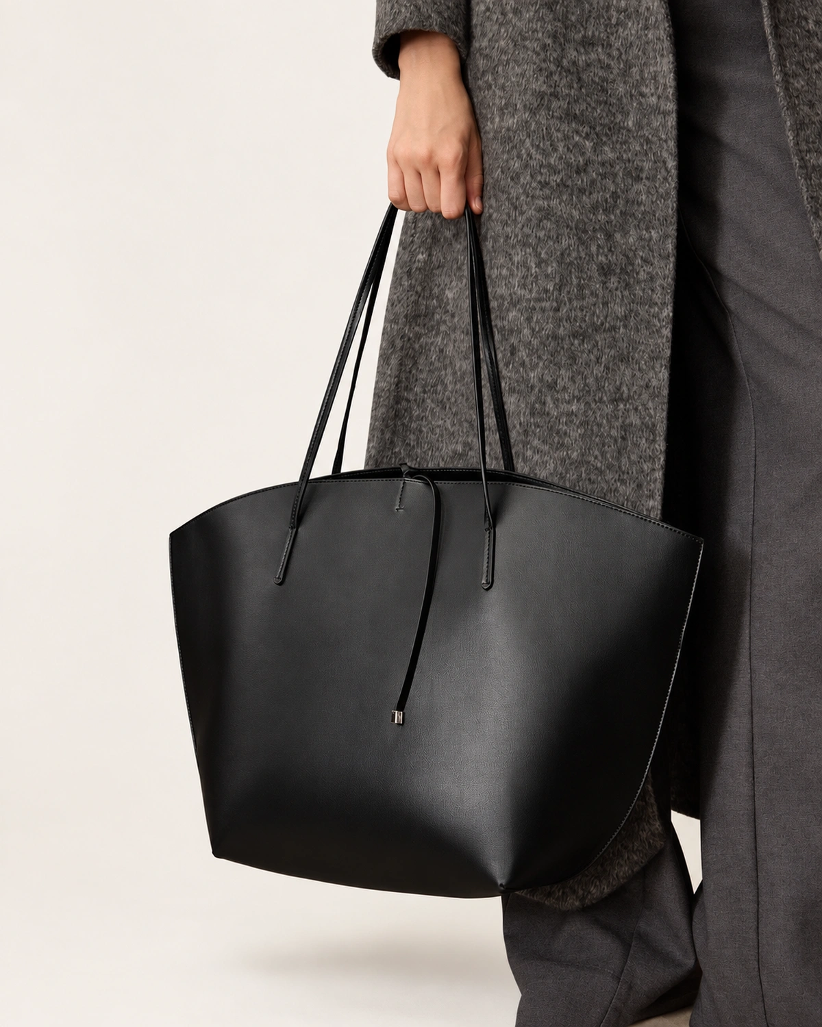 Plano detalhe da mão da modelo segurando a alça da bolsa tote preta ao lado da calça de alfaiataria cinza.