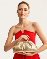 Modelo feminina em plano médio com vestido de festa vermelho segurando a clutch drapeada dourada metalizada na cintura.