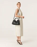 Modelo feminina de corpo inteiro vestindo vestido claro e sandálias de salto segurando a bolsa tote preta a tiracolo.