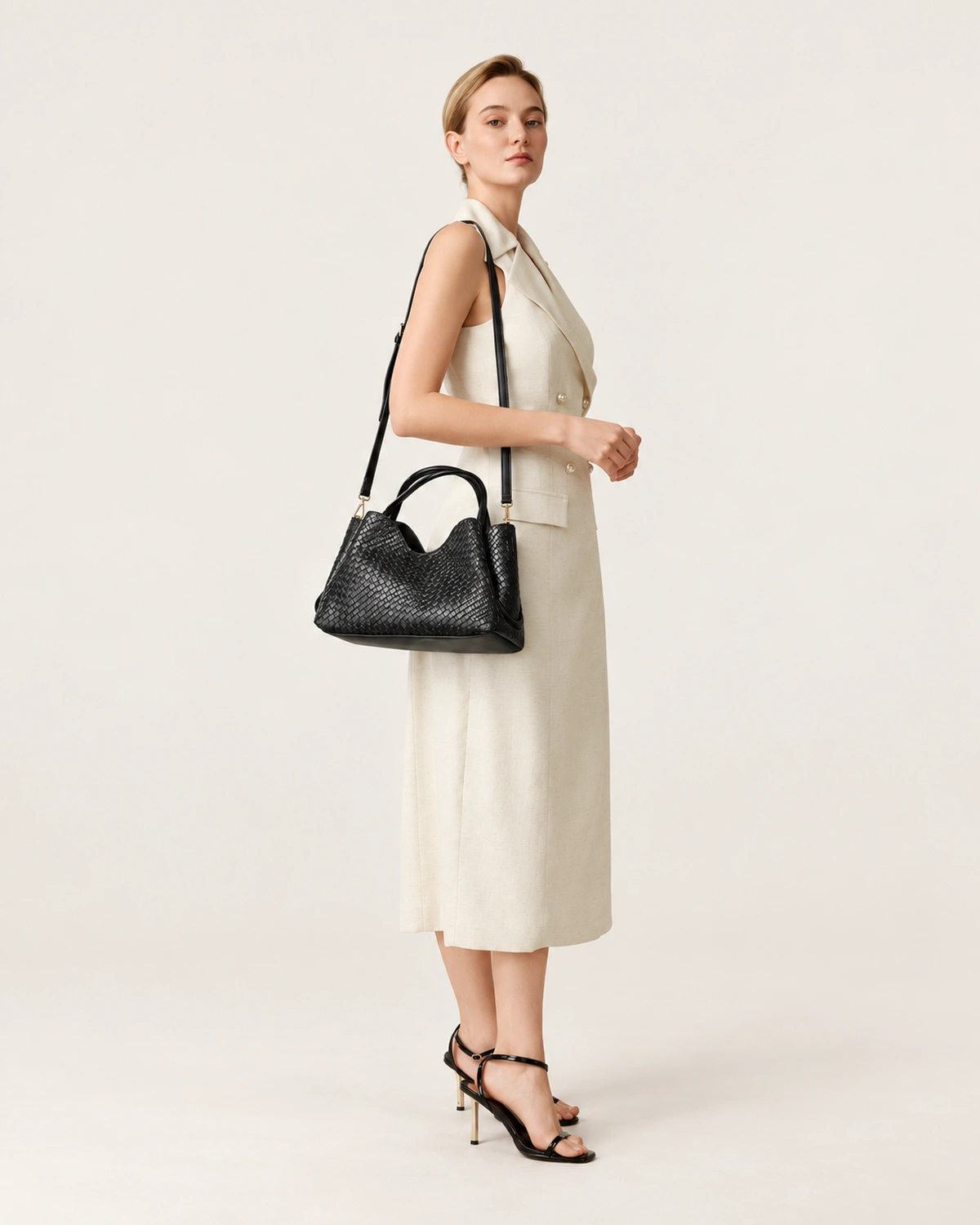 Modelo feminina de corpo inteiro vestindo vestido claro e sandálias de salto segurando a bolsa tote preta a tiracolo.
