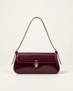 Vista frontal isolada da bolsa tiracolo flap bordeaux com fecho metálico prateado assentada no solo do estúdio mineral.