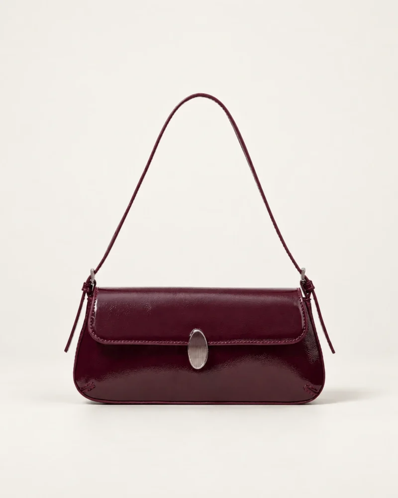 Vista frontal isolada da bolsa tiracolo flap bordeaux com fecho metálico prateado assentada no solo do estúdio mineral.