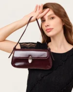 Modelo feminina em plano editorial segurando a bolsa tiracolo flap em couro acetinado bordeaux no estúdio mineral.