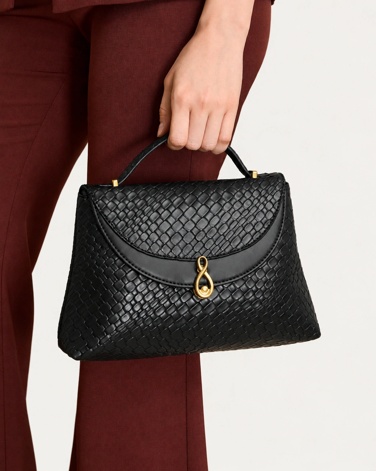 Detalhe da mão da modelo segurando a alça da Bolsa Satchel Estruturada preta contra look alfaiataria bordô.