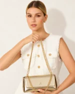 Plano médio da modelo loira feminina segurando a bolsa tiracolo estruturada dourada à frente do tronco com conjunto em tweed.