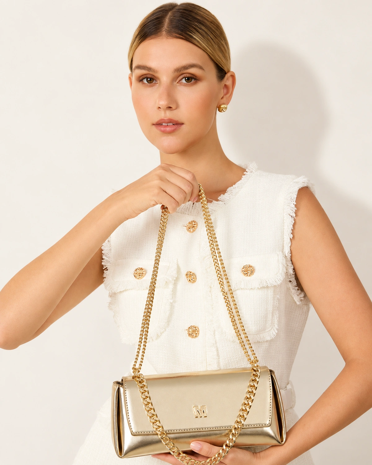Plano médio da modelo loira feminina segurando a bolsa tiracolo estruturada dourada à frente do tronco com conjunto em tweed.
