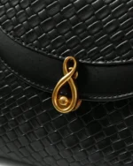 Close macro do fecho metálico dourado escultural e textura tressê em relevo da Bolsa Satchel preta.