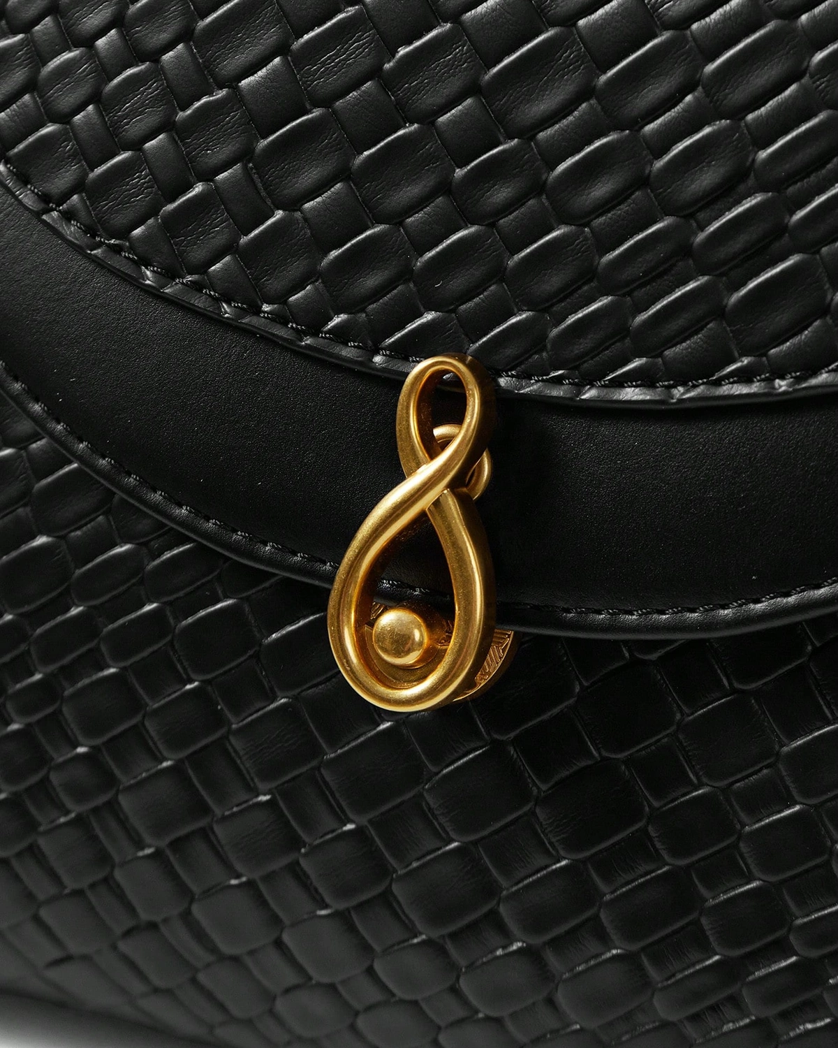 Close macro do fecho metálico dourado escultural e textura tressê em relevo da Bolsa Satchel preta.