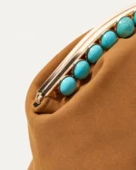 Close-up diagonal da armação dourada com contas turquesa e acabamento aveludado da bolsa clutch marrom