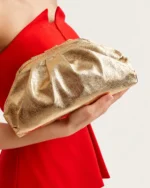 Plano detalhe das mãos da modelo segurando a clutch drapeada dourada metalizada contra o vestido vermelho.