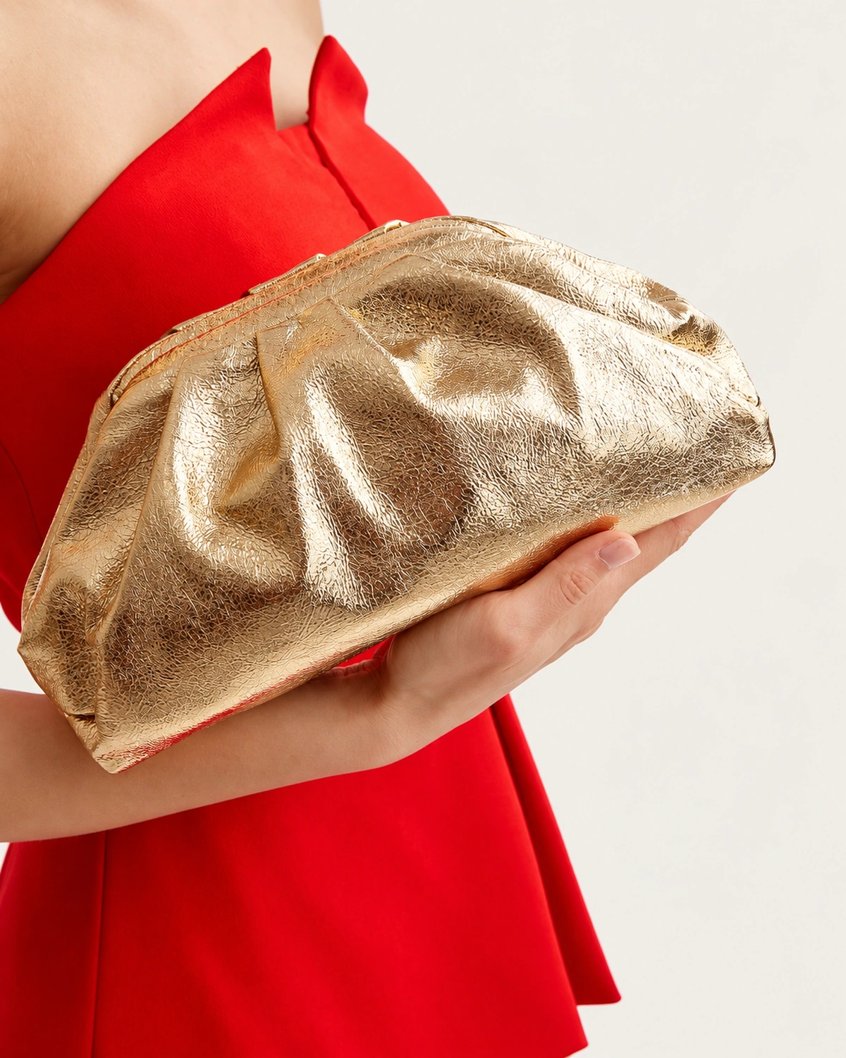 Plano detalhe das mãos da modelo segurando a clutch drapeada dourada metalizada contra o vestido vermelho.