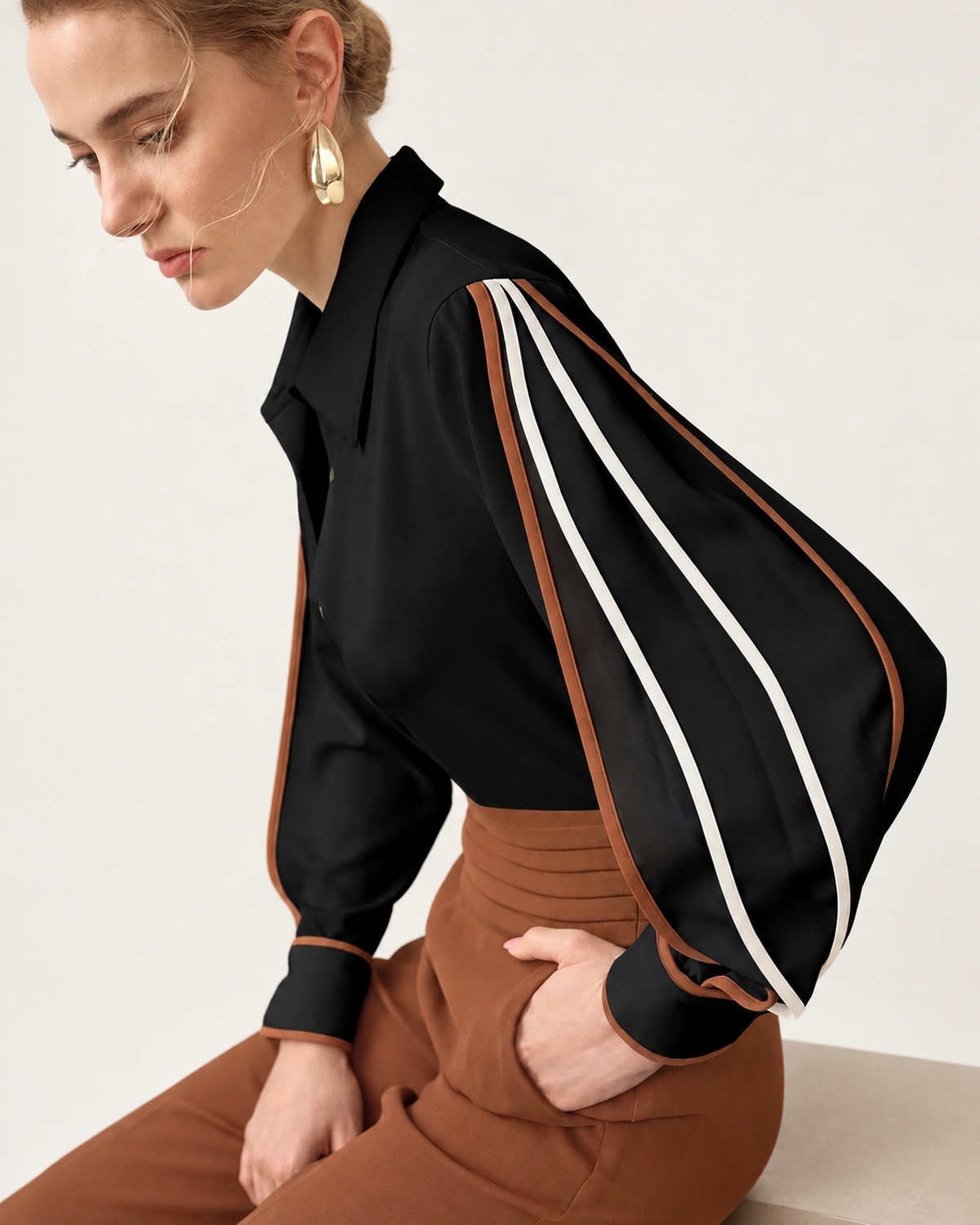 Modelo sentada de perfil vestindo camisa preta com vivos contrastantes e calça pantalona marrom
