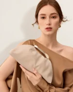 Bolsa Satchel Drapeada com Alça Escultural Metálica em tom areia com alça escultural metálica.